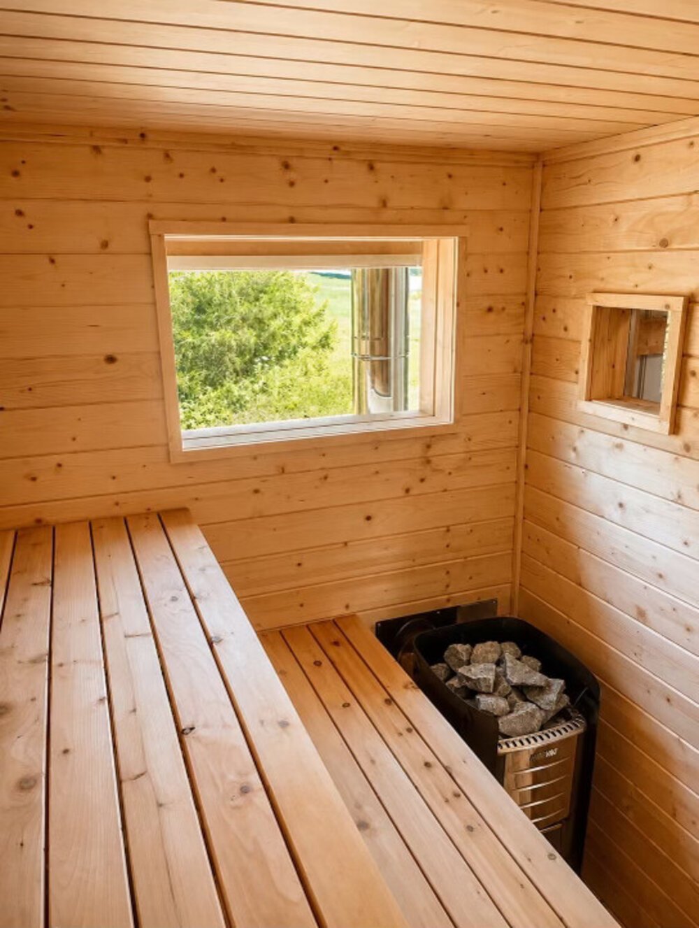 Interior of a cedar sauna with a window view and a wood-fired heater topped with stones